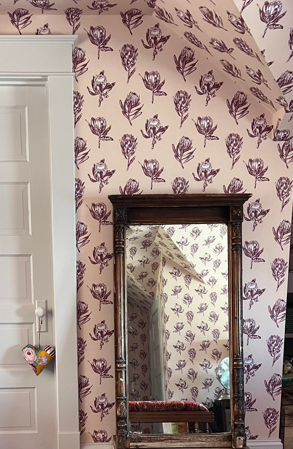 A vintage-style wooden bench with a vibrant red, orange, and purple paisley upholstered seat cushion, positioned in front of a tall rectangular mirror on a beige carpeted floor. The background features a white paneled door on the left and a wall covered in pink floral wallpaper with a matching floral pattern on the mirror&