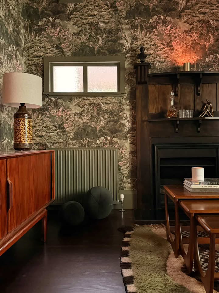 Arcadia wallpaper, premium wallpaper. A styled interior room featuring a dark wood mid-century sideboard with a patterned table lamp, a window, a green radiator, two dark poufs, a dark wood fireplace mantel, 