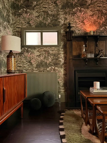 Arcadia wallpaper, premium wallpaper. A styled interior room featuring a dark wood mid-century sideboard with a patterned table lamp, a window, a green radiator, two dark poufs, a dark wood fireplace mantel, 