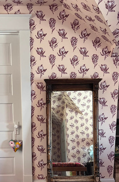 A vintage-style wooden bench with a vibrant red, orange, and purple paisley upholstered seat cushion, positioned in front of a tall rectangular mirror on a beige carpeted floor. The background features a white paneled door on the left and a wall covered in pink floral wallpaper with a matching floral pattern on the mirror&