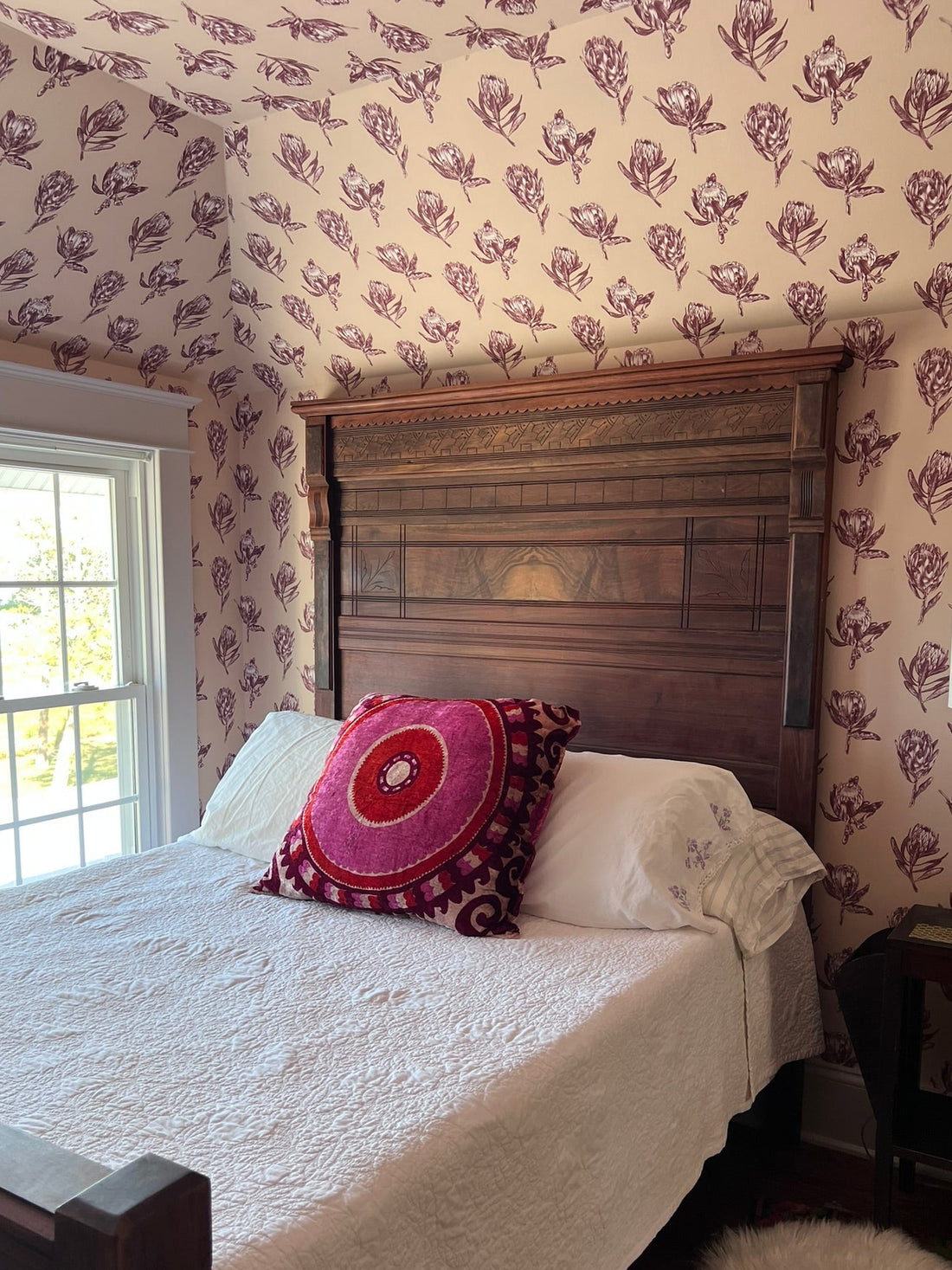 A bed dressed in white textured bedding with a round pink and red mandala-patterned accent pillow, set against a dark wood headboard in a bedroom with floral wallpaper.