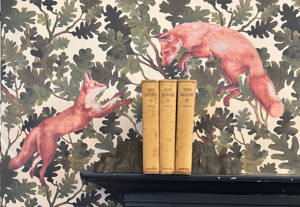 Two fabric fox decals leaping across a green oak-leaf wallpaper backdrop, displayed above a black cast-iron fireplace with three vintage 'Home Mechanic' books between antique tree-stump bookends.