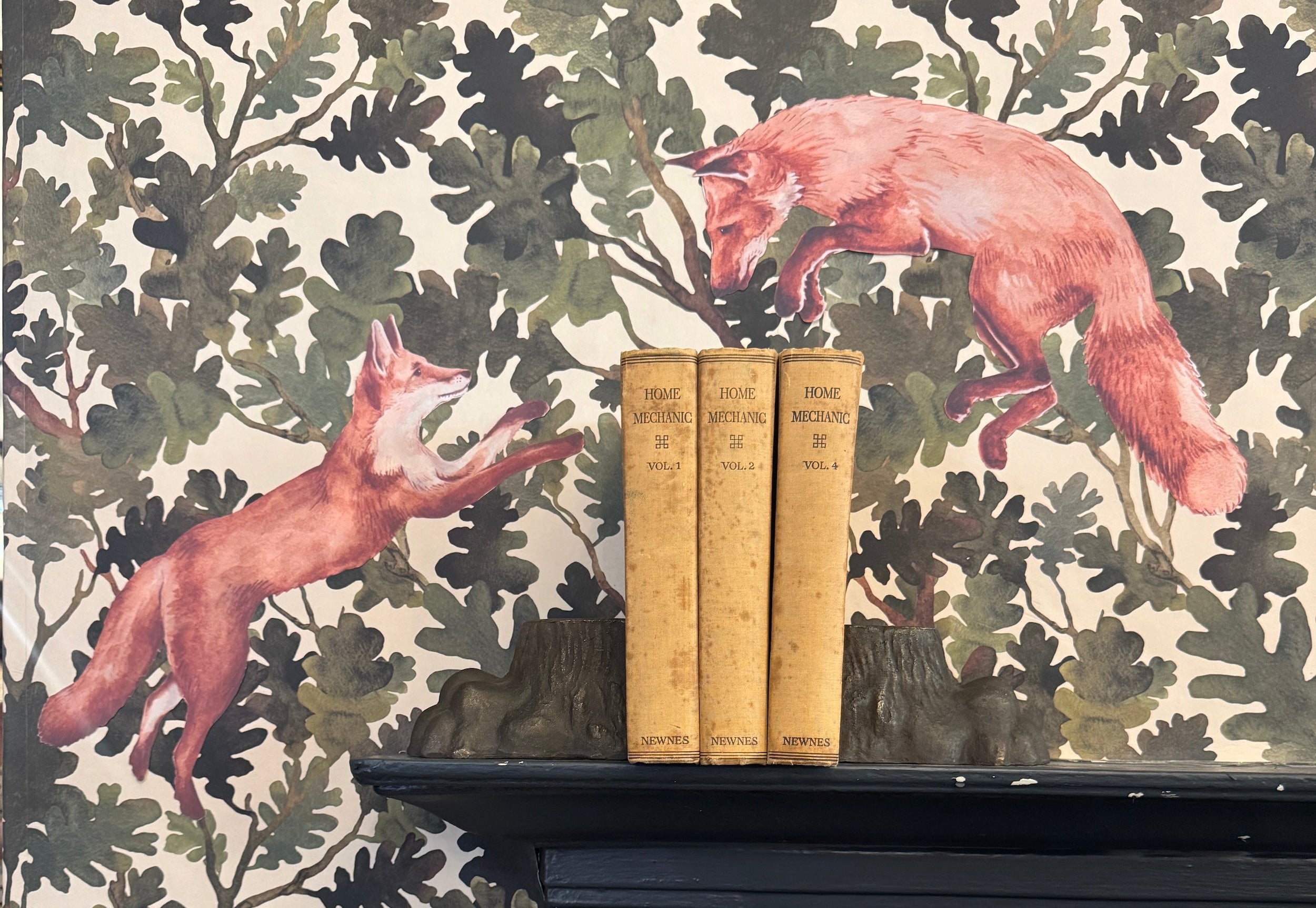 Two fabric fox decals leaping across a green oak-leaf wallpaper backdrop, displayed above a black cast-iron fireplace with three vintage 'Home Mechanic' books between antique tree-stump bookends.