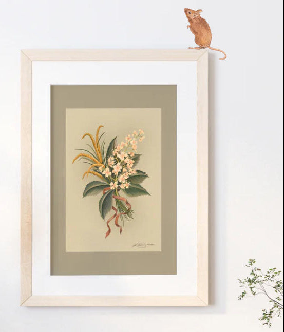 A framed botanical art print featuring a vintage illustration of a flower bouquet with green leaves and cream blossoms, mounted on a light beige wall with a small mouse figurine perched on the top of the frame.