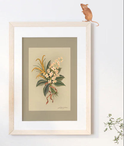 A framed botanical art print featuring a vintage illustration of a flower bouquet with green leaves and cream blossoms, mounted on a light beige wall with a small mouse figurine perched on the top of the frame.